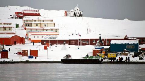 A series of single storey building coloured brown and orange stand in the snow comprising Chile's Presidente Eduardo Frei military base next to the Antarctic Ocean. People can be seen working along the shoreline and there is an open-topped boat in the water. 