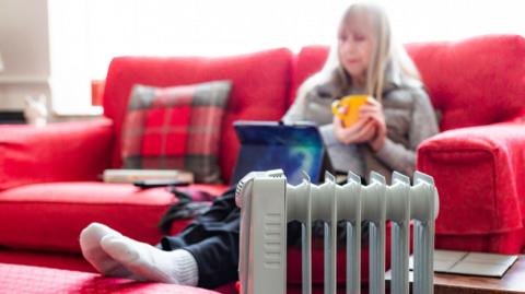 An older woman sits on a couch with a cup between her hands. She has her feet on a stool next to a heater