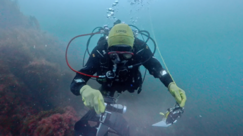 Kevin McIlwee is wearing a black wetsuit, mask, regulator, gloves and a bright yellow hood. He's swimming close to a rocky seabed covered with marine growth and is holding camera equipment in one hand while following a thin guideline that extends upward through the water. Air bubbles rise above the him toward the surface, and the surrounding water is blue-green with moderate visibility.