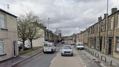 Rows of terraced houses line Rochdale Road on a cloudy day. There are two cars and a white van on the road in this Google Maps image. 