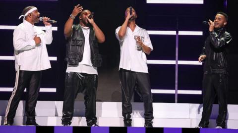 Boyband JLS members standing on stage performing a song singing into microphones. They are all dressed in black and white outfits, with one member wearing a white band around his head. The members are stood in front of a black screen, with white stripes on it and a white step at the bottom.