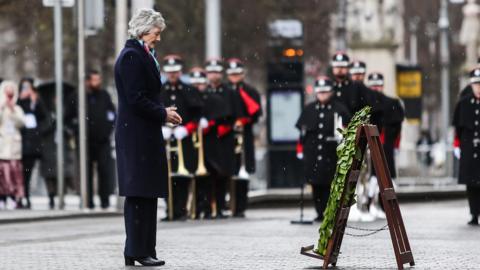 A woman with grey short hair stands facing a green wreath - she is wearing a navy coat with black trousers and shoes. it is raining.