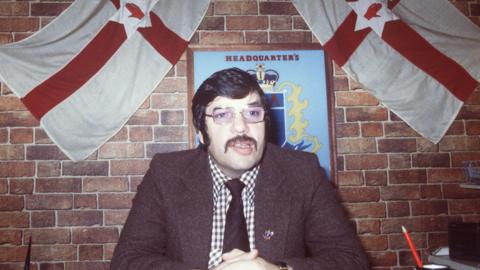 A man in a brown tweed jacket, gingham shirt, brown tie and glasses. He has dark hair combed neatly and a long brown moustache. Behind him are two ulster flags and a blue plaque which says 'UDA Headquarters'.