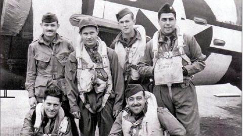 A black and white photo of US aircrew dressed in caps and flying attire posing for the camera in front of an aircraft.