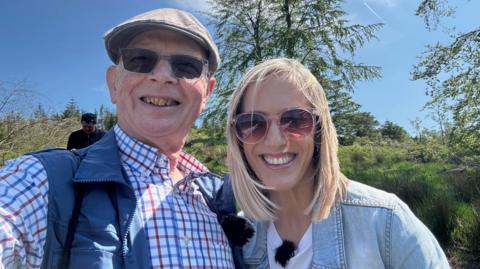 Kay Crewdson is standing next to Roger Hordley in Macclesfield Forest. They are both smiling at the camera and are wearing sunglasses. Kay is wearing a denim jacket and white T-shirt and Roger is wearing a chequered shirt and blue gilet.