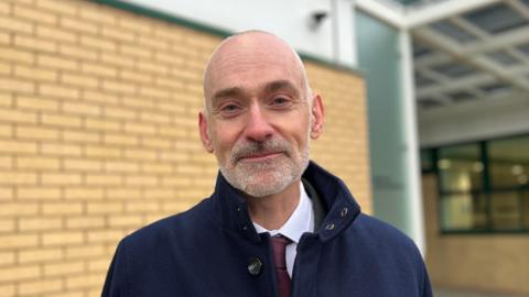 Professor Peter Bradley is a man with short grey hair and a grey beard. He is looking into the camera. He is standing outside a pale brick building.