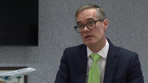 Chris Woodroofe, wearing a dark blue suit jacket and lime green tie over a white shirt, speaks to the camera with a model Aer Lingus aircraft visible in the background