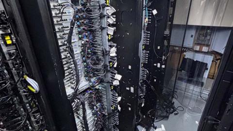 A row of computer servers, tall stacked black computer cabinets with multiple trays and wire connections
