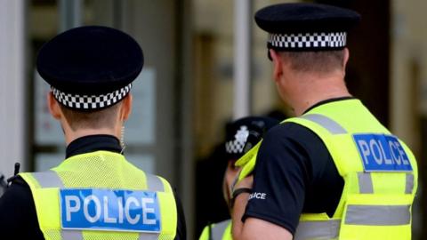 The backs of two male police officers stood talking to a female police officer. You can only see part of her face. 