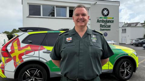 John Atkins has short grey hair and is wearing a green short sleeved uniform shirt with a Guernsey flag, the Ambulance and Rescue crest and his name badge on it, is smiling at the camera. He's standing in front of an ambulance branded Volvo with wide green and luminous yellow stripes on it and the Guernsey flag across the rear panel of the driver's side. On the building in the background you can see the Ambulance and Rescue Guernsey logo on the wall.