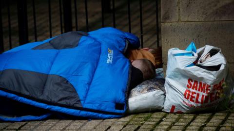 Two people lie on a street corner in a blue sleeping bag. Behind them, there is a plastic bag. 