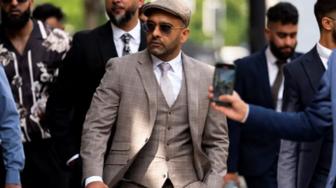 Akhmed is wearing a beige patterned suit with a waistcoat, white shirt, flat cap and sunglasses. He is walking on a street and has several people flanking him.