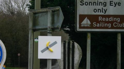 A banana duct-taped onto a white board is affixed to the pole of a street sign on a mini roundabout