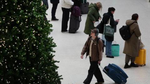People gather in train station with suitcases, a Christmas tree with lights on it is in the foreground