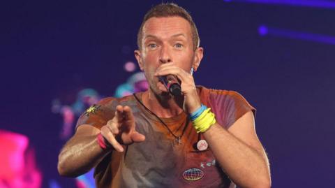  Coldplay's Chris Martin performing on stage at Wembley Stadium, north London, in 2022. He has short brown hair and stubble and is holding the microphone up to his mouth with his left hand. His right hand is outstretched. He is wearing a reddish t-shirt