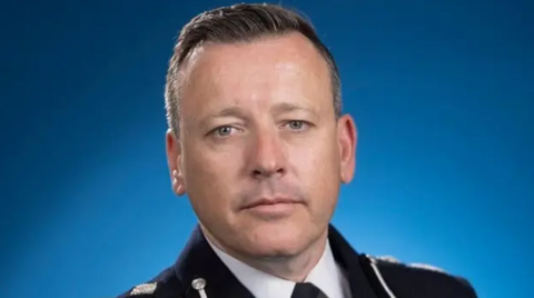 A close-up photo of the acting chief constable of West Midlands Police. He wears a black police uniform and stands in front of a blue background, looking into the camera.