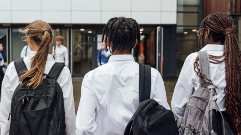 A stock image of the backs of three secondary school age students walking into a school entrance.