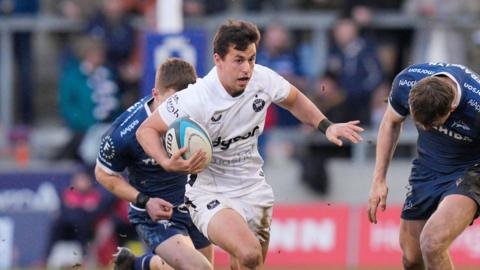 Neil Le Roux runs with the ball in his right arm as a Sale player chases behind him and another stands with his head down to his left
