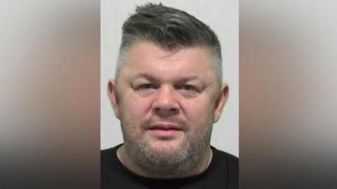 Anthony Craggs' mugshot. He has a grey stubbly beard with short dark hair swept over to the right and shaved grey sides.