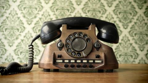 A vintage rotary dial telephone sits on a wooden surface. The phone is made of dark, worn metal with a black handset resting on top. A coiled cord runs from the left side of the base. Behind it is patterned green wallpaper with an ornate, repeating design.