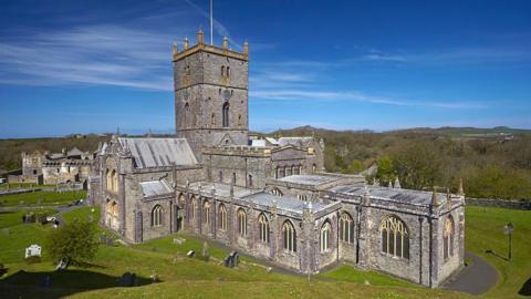 An exterior view of St Davids Cathedral. A large stone building with a square tower, it is surrounded by a grassy churchyard.