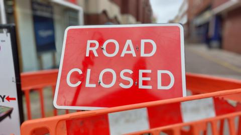 A close up photo of a red sign with white writing, saying ROAD CLOSED. It is set among orange barriers, with a blurry street behind it.
