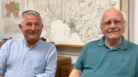 Richard sits on the left in a light blue shirt. Reg is on his right, he is wearing a green polo shirt and has glasses. They both have white hair.