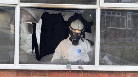 A man in an all white hazard suite and wearing a mask with a long snout over a hard hat with a lamp at the front. He is pictured behind a broken window. the room inside looks very dark