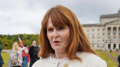 Caoimhe Archibald speaking into a microphone. She is wearing a cream jacket with gold buttons. She has long red hair. She is standing outside Stormont parliament buildings.