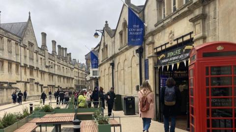 A street with old buildings on either side stretched into the distance and curves to the right. There are various people dotted around, mostly wearing wintry clothing. Two blue flags jutting out of the wall of a building on the right side of the road read 'The Covered Market est. 1974'. The entrance to the market sits under a banner with 'Open Late' written on it. Two people with their backs turned, both wearing backpacks, appear to be making their way inside. 
