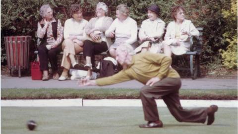 Man takes a bowling shot on a green, with six elderly ladies sitting together on a bench in the background