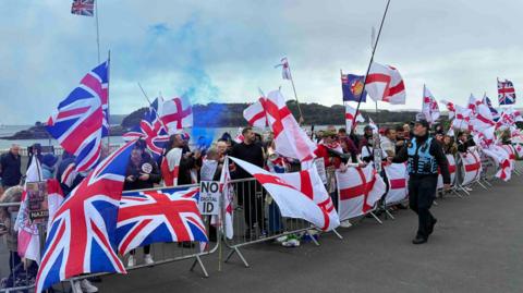 A group of protestors on Plymouth Hoe behind railings waving the Union Jack and St George's flags. A blue flare has been set off and police watch the scene.