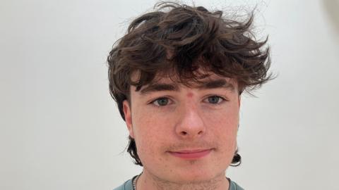 A young man with curly brown hair, looking at the camera. He is pictured from the shoulders up. There is a light grey wall behind him. 