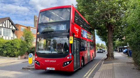 A red London bus is driving along a tree-lined suburban street. There are mid 20th Century houses on both sides of the road. 