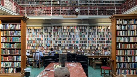 The inside of the library with cases and cases of books. People are reading in the background. It is on two floors.