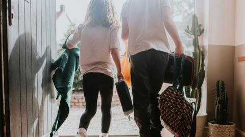 Stock picture of the back of two children leaving a house. They are wearing school uniform and carrying bags.