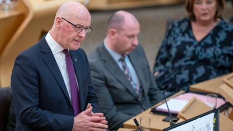 John Swinney, a bald man with glasses, is wearing a blue jacket, white shirt and purple tie. He is standing up to answer a questions with has hands together in front of him. He has a male and female colleague sitting beyond him