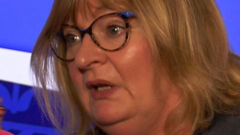 A woman speaking into a studio microphone during a broadcast, seated against a blue backdrop, wearing a dark shirt.