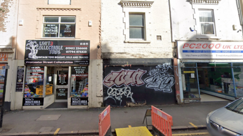 The building sits in between two businesses. The top half of the facade is white brick, with a window and an air vent. The lower half of the building is covered by black boards covered in graffiti that have been put in place. One business on the left looks to be a collectables toy shop and on the right a printing or printer-related store.