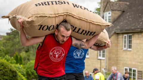 A young man with a red T-shirt and 'Tetbury Woolsack Races' written across it carries a large sack up a hill as part of the Tetbury woolsack races. Behind him is another man with a blue T-shirt with the same logo.