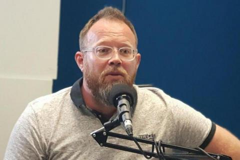 Neil Maggs with a blondish-brown beard and translucent glasses is sitting in a studio, wearing a grey T-shirt with black trim. He is speaking into a microphone. Behind him is a blue and white background.