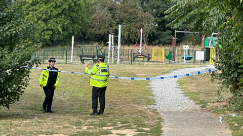 Two police officers standing in a park talking to each other. Police tape can be seen dangling between two trees.