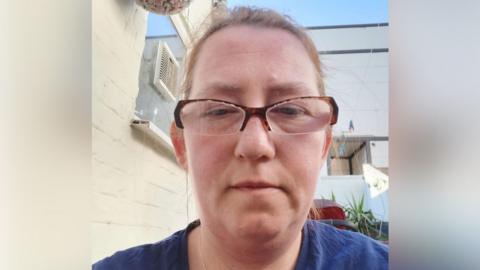 Marta Bednarczyk with short brown hair and brown glasses, looking at the camera and wearing a blue top. She is in front of a white building.