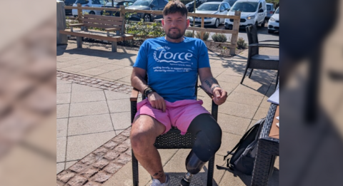 The picture shows a person sitting outdoors on a chair, wearing a blue t-shirt with the word "force". They are also wearing pink shorts and white trainers. The person has a prosthetic leg.