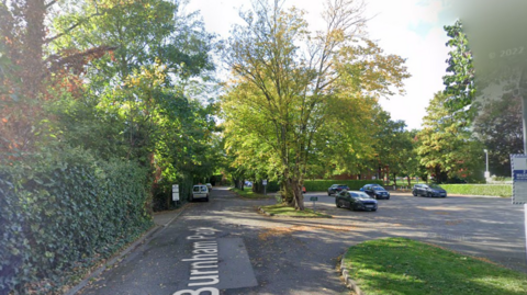 A screenshot from Google street view showing a car park and a road lined with trees on a sunny day.