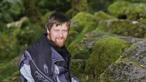 Ben Rowe, with a beard, is wearing a motorcycling jacket and sitting on rocks covered in moss in his former Facebook profile picture