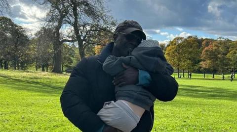 A man in a black puffer jacket and dark peach trousers is hugging a child in grey tracksuits, wellington boots, a hoody and a wooly hat. They are standing in a park