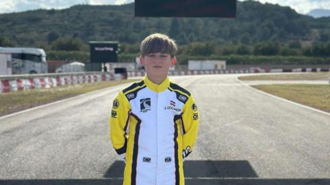 Jack Collinson, a boy with short blond hair wearing a yellow and white one-piece racing suit, is standing with the empty track in Valencia behind him. 