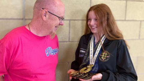 Alan wearing a pink T-shirt standing next to Lana, wearing an England branded black or navy blue jumper, while she shows him her medals around her neck- smiling at each other.