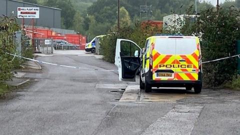 A police van is pictured in an industrial estate, with police cordon tape spanning the width of the road